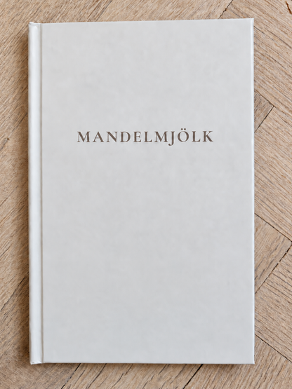 Mandelmjölk anteckningsbok med ljust omslag och mörkbrun titel på ljus fiskbensparkett